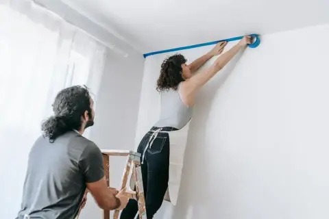 Para przygotowuje się do malowania ścian. Kobieta na drabinie przykleja taśmę malarską, mężczyzna ją asekuruje.